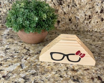 May include: A wooden eyeglasses holder with a black outline of glasses and a red glitter bow. The holder is triangular and sits on a speckled countertop. A small potted plant is in the background.
