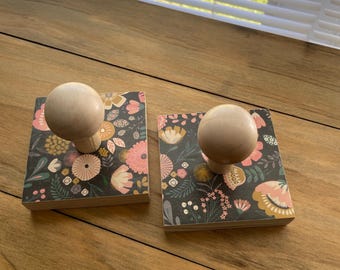 May include: Two square wooden coasters with a floral design in pink, yellow, and white on a dark background. Each coaster has a round, light-colored wooden knob on top. The coasters are displayed on a wooden surface.