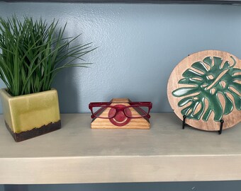 May include: A decorative shelf arrangement featuring a green potted plant, a wooden eyeglass holder with red glasses, and a wooden plaque with a green leaf design. The items are arranged on a light-colored shelf against a blue wall.