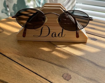 May include: A wooden sunglasses stand with the word "Dad" engraved on it. A pair of round, brown sunglasses are resting on the stand.