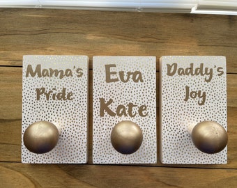 May include: Three rectangular wooden plaques with gold-colored knobs. Each plaque is white with gold polka dots and gold lettering. The plaques read "Mama's Pride", "Eva Kate", and "Daddy's Joy".