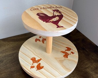 May include: A two-tier wooden serving stand with a rooster design. The top tier has the text "Cock a doodle doo" and a red rooster. The bottom tier has a brown rooster and several smaller brown chicks.