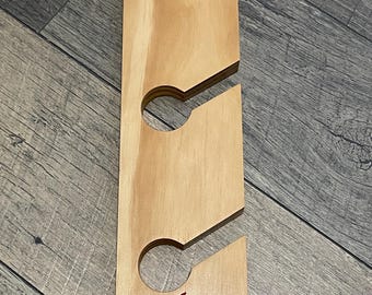 May include: A wooden wine glass holder with three circular cutouts and angled slots. The natural wood-toned piece features a red skull and flame logo at the bottom. The holder is designed to hold wine glasses.