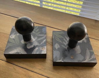 May include: Two dark wooden bookends with a floral design. Each bookend has a square base with a dark floral pattern in shades of blue, gray, and white. A dark wooden sphere is attached to the top of each base.