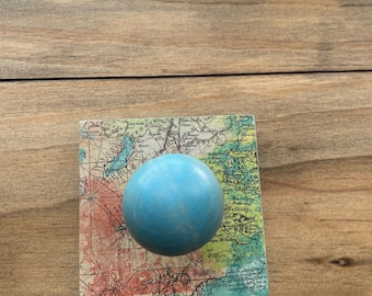 May include: A square coaster with a vintage map design and a light blue sphere on top. The coaster has a colorful map print with red, green, and blue hues. The sphere is a solid, light blue color.