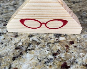 May include: A wooden eyeglass holder in a pyramid shape. The holder is made of light-colored wood and features a red outline of eyeglasses on the front. The top of the holder has grooves for holding glasses.