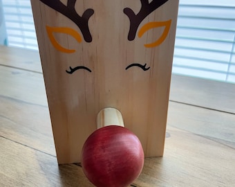 May include: A wooden reindeer-themed decoration with a red nose and brown antlers. The face features orange and black accents, including closed eyes. The wooden block is rectangular and stands upright.