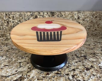 May include: A wooden cupcake stand with a round, light-colored top featuring a cupcake design with red, white, and black details. The base is black and round. The stand is approximately 6 inches in diameter.