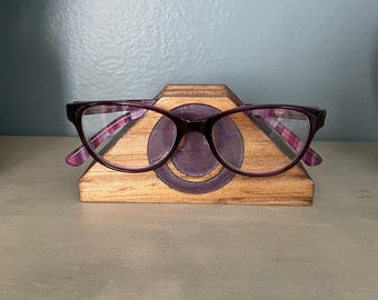 May include: A pair of purple-framed eyeglasses rests on a wooden eyeglasses holder. The glasses have a rectangular shape with a plaid pattern on the temples. The holder is made of wood and has a circular design.