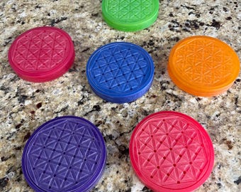 May include: Six colorful, round coasters in shades of red, green, blue, orange, and purple. Each coaster features a geometric pattern of intersecting lines and shapes. The coasters are arranged on a speckled surface.