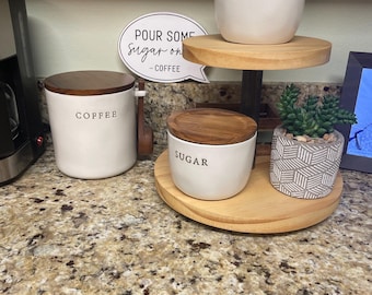 May include: A tiered wooden tray holds a cream pitcher labeled "CREAM" and a small succulent in a geometric pot. Below, a jar labeled "SUGAR" and a "COFFEE" jar with a wooden lid sit on the tray. A sign reads "POUR SOME Sugar on - COFFEE."