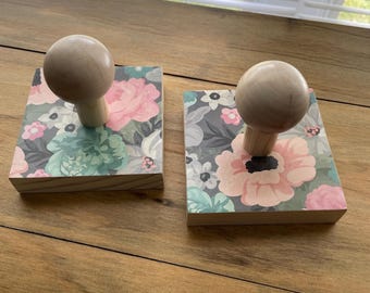 May include: Two wooden coasters with floral patterns and a light-colored wooden knob on top. The coasters feature pink, blue, and gray flowers on a white background. The coasters are displayed on a wooden surface.