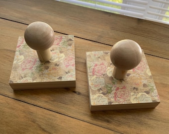 May include: Two wooden stamps with a floral design. Each stamp has a square base with a light-colored floral pattern and a round wooden handle. The stamps are displayed on a wooden surface.