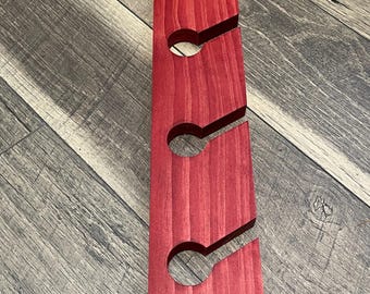 May include: A vertical, red-stained wooden wine rack with three circular openings and angled slots to hold wine bottles. The rack is made of solid wood and has a smooth finish. The design is simple and functional.