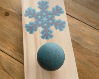 May include: A wooden snowflake ornament with a teal blue knob. The snowflake is painted in gray and teal blue.
