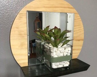 May include: A round wooden mirror with a rectangular mirror insert and a black shelf. A square glass vase with white stones and green-tipped plants sits on the shelf. The mirror's wooden frame is light-colored.