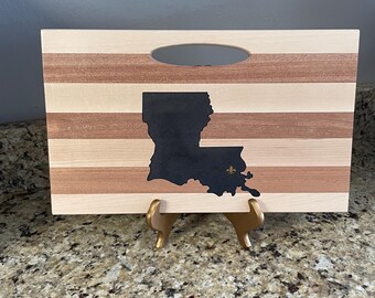 May include: Wooden cutting board with a handle, featuring horizontal stripes of light and dark wood. A black silhouette of the state of Louisiana is centered, with a gold fleur-de-lis. The board is displayed on a gold stand.