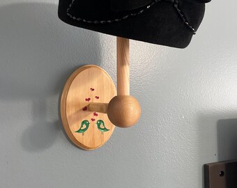 May include: A black cloche hat with a large bow and decorative trim is displayed on a wooden hat rack. The rack features a round wooden peg and a decorative wooden plaque with painted green birds and red hearts.