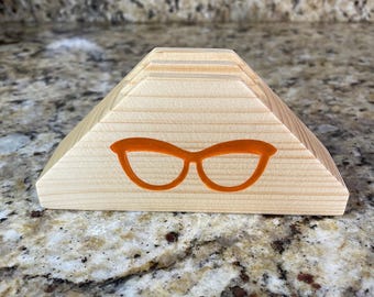 May include: A wooden eyeglass holder in a pyramid shape. The holder is made of light-colored wood and features an orange outline of eyeglasses on the front. The top has grooves for holding glasses. The holder is on a speckled surface.