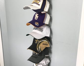 May include: A collection of baseball caps in various colors and designs, displayed on a wooden rack. The caps feature team logos and text, including "LSU", "Saints", and "Champions". The caps are arranged vertically against a light blue wall.