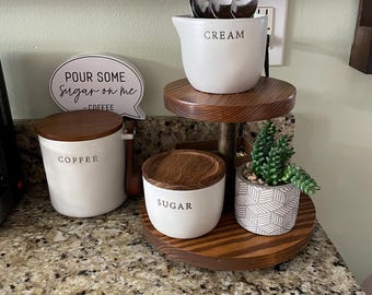 May include: A tiered wooden stand with a white ceramic creamer labeled "CREAM" and a white ceramic sugar dispenser labeled "SUGAR". The stand also features a white ceramic coffee container labeled "COFFEE" and a small potted succulent plant. A white sign with black text reads "POUR SOME SUGAR ON ME - COFFEE".