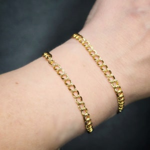 Puede incluir: Dos pulseras de cadena dorada en la muñeca de una persona. Las pulseras están hechas de una cadena gruesa y enlazada.