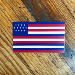 Puede incluir: Una pegatina rectangular con un diseño estilizado de la bandera estadounidense. La bandera tiene un campo azul con 13 estrellas blancas y rayas rojas, blancas y azules. La pegatina se muestra sobre una superficie de madera.