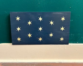 May include: A dark blue rectangular sign with 13 white five-pointed stars arranged in a pattern. The sign is a representation of the original American flag.
