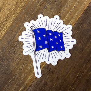 May include: A blue and white sticker with a stylized illustration of a flag with white stars on a blue background. The flag is waving in the wind and has a white pole. The sticker has a white background with a blue sunburst design around the flag.