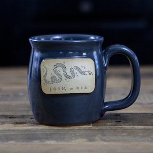 May include: A dark blue ceramic mug with a white handle. The mug features a vintage illustration of a snake with the text "JOIN, or DIE." and the initials of the original 13 colonies.