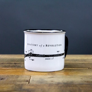 May include: White ceramic mug with a black rim and handle. The mug features a black and white illustration of a gun with the text "Anatomy of a Revolution" and labels for the parts of the gun.