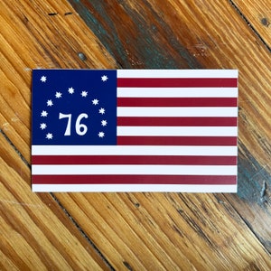 May include: Rectangular sticker featuring a stylized American flag design. The flag has a blue canton with white stars surrounding the number "76". The flag's stripes are red and white. The sticker is placed on a wooden surface.