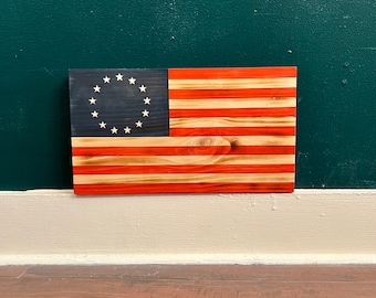 Könnte beinhalten: Eine amerikanische Holzflagge mit 13 roten und weißen Streifen und einem blauen Feld mit 13 weißen Sternen in einem Kreis. Die Flagge ist an einer Wand montiert.