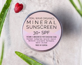 Op de afbeelding: Een ronde, zilveren blik van Tidal Wave Organics Mineral Sunscreen 30+ SPF, met tekstdetails. De blik is omgeven door aloë vera bladeren en rode frambozen. Het product is veganistisch en ongeparfumeerd, gemaakt in Florida.
