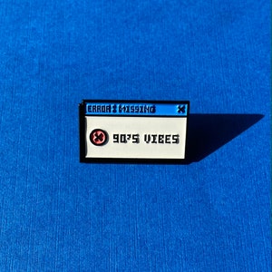 Könnte beinhalten: Rechteckiger Emaille-Pin mit schwarzem Rand, mit einem pixeligen Design, das an eine Computerfehlermeldung erinnert. Der obere Teil ist blau mit dem Text "ERROR: MISSING". Der untere Teil ist weiß mit "90'S VIBES" und einem rot-schwarzen Symbol.