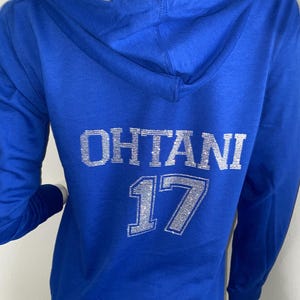 May include: A royal blue hooded sweatshirt with the name "OHTANI" and the number "17" in silver glitter lettering on the back. The hood is lined with a light gray fabric. The sweatshirt has long sleeves.