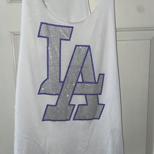 May include: White vest top featuring the letters "LA" in silver rhinestones with a blue outline. The vest has a racerback design and is hanging on a wooden hanger.