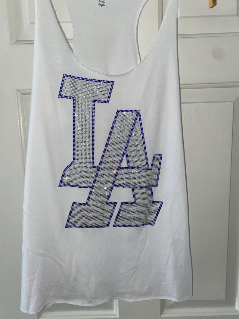 May include: White vest top featuring the letters "LA" in silver rhinestones with a blue outline. The vest has a racerback design and is hanging on a wooden hanger.