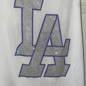 May include: White vest top with a sparkling "LA" design. The letters are outlined in blue rhinestones and filled with silver rhinestones. A casual, sporty top.