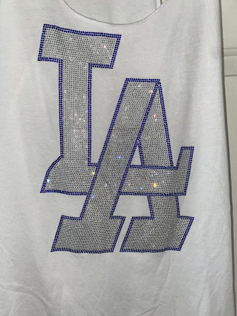 May include: White vest top with a sparkling "LA" design. The letters are outlined in blue rhinestones and filled with silver rhinestones. A casual, sporty top.