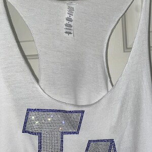 May include: White vest top featuring the letters "LA" in blue and silver rhinestones. The vest has a racerback design and is hanging on a wooden hanger. The top is made of a soft, lightweight material.