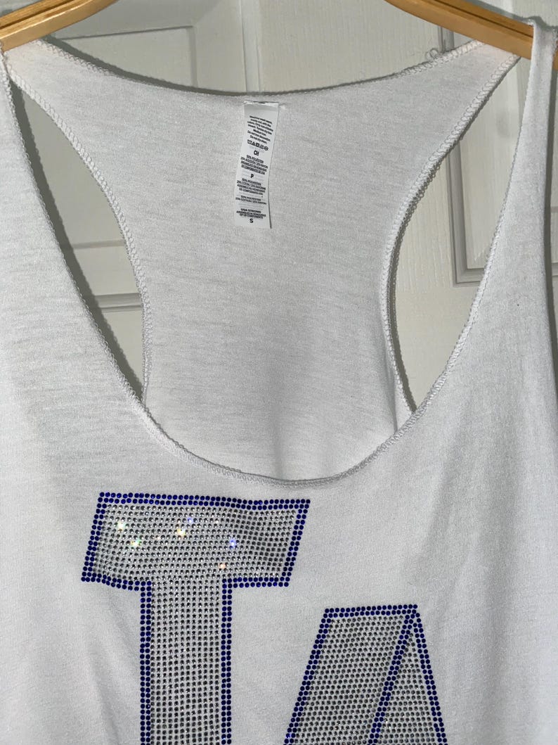 May include: White vest top featuring the letters "LA" in blue and silver rhinestones. The vest has a racerback design and is hanging on a wooden hanger. The top is made of a soft, lightweight material.