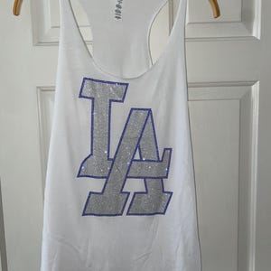 May include: White vest top with a racerback design. The front displays the letters "LA" in silver with blue rhinestone accents. The vest top is hanging on a wooden hanger.