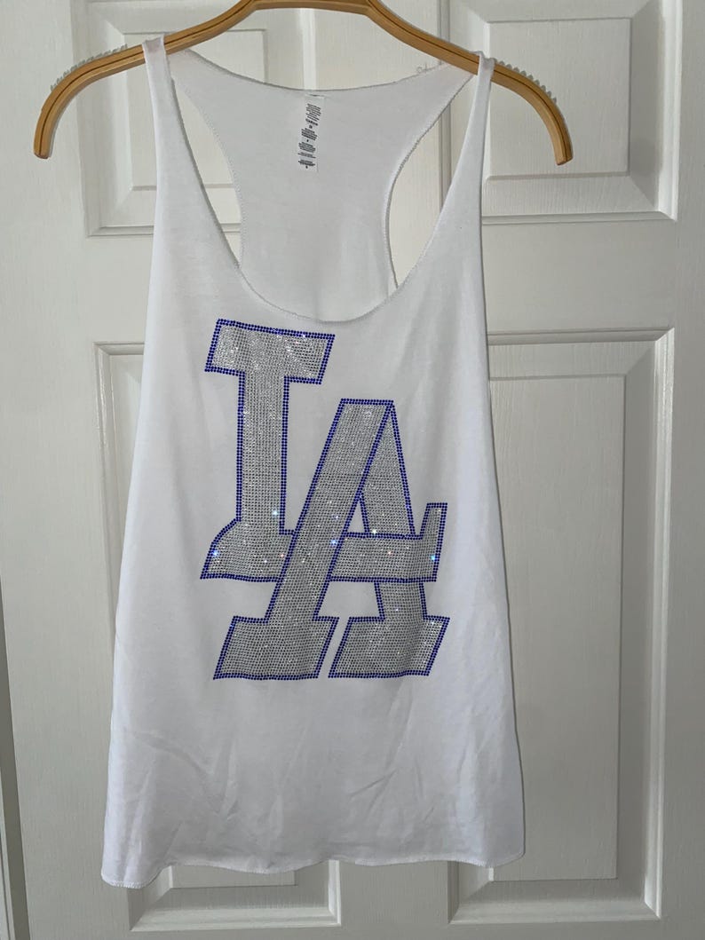 May include: White vest top with a racerback design. The front displays the letters "LA" in silver with blue rhinestone accents. The vest top is hanging on a wooden hanger.