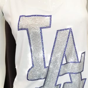 May include: White vest top featuring the letters "LA" in silver rhinestones with a blue outline. The sleeveless top has a V-neck. The garment is displayed on a mannequin.