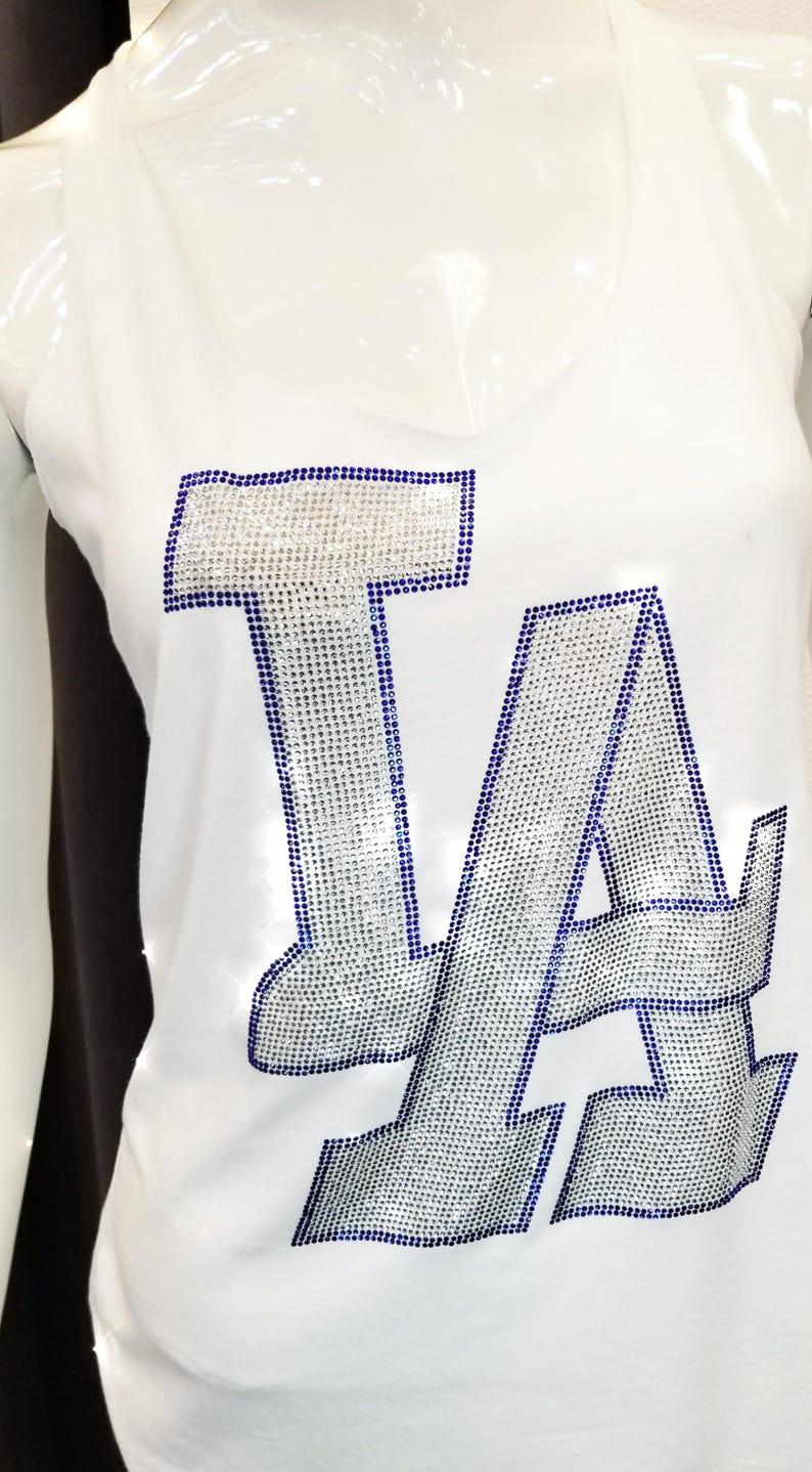 May include: White vest top featuring the letters "LA" in silver rhinestones with a blue outline. The sleeveless top has a V-neck. The garment is displayed on a mannequin.