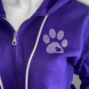 May include: A purple zip-up hoodie with a rhinestone paw print design on the chest. The zipper is silver, and a rhinestone trim runs along the zipper and hood. The hoodie is displayed on a white mannequin.