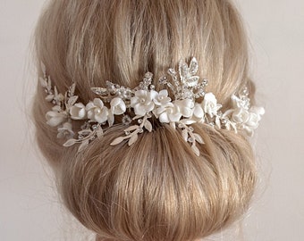 May include: A white bridal hair comb with delicate white flowers and silver leaves. The comb is worn in a woman's blonde hair, styled in a low bun.