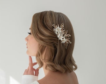 May include: A delicate hair comb featuring white fabric flowers, gold-toned accents, and small pearl-like beads. The comb is set in a woman's hair, adding a touch of elegance. Perfect for weddings or special occasions.