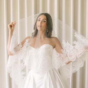 May include: A bride in a white strapless wedding dress and veil. The veil and dress feature intricate lace detailing. The bride is holding the veil open, showcasing the design. The background is a neutral, cream-colored curtain.